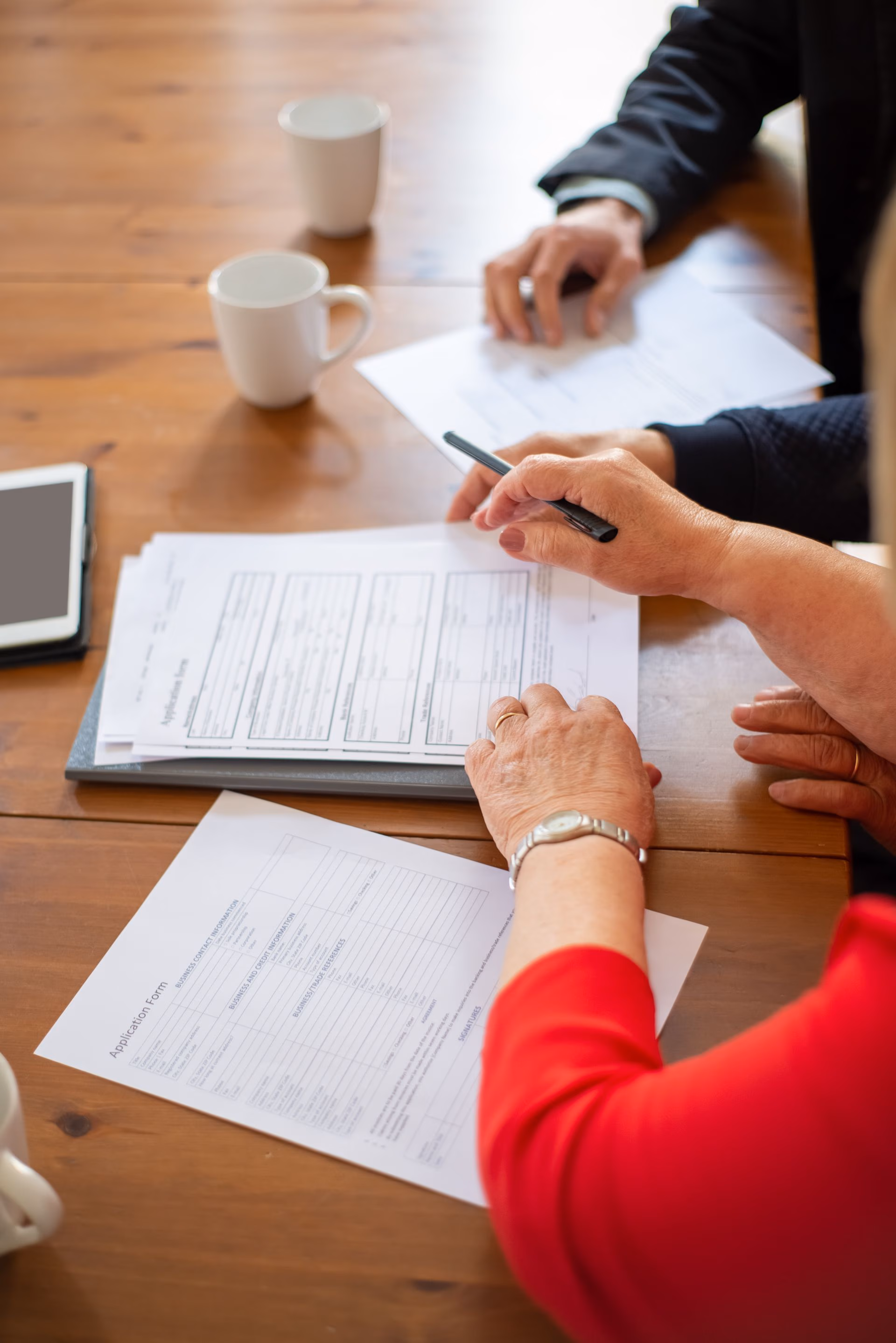 Two people reviewing and filling out forms at a wooden table with cups and a tablet nearby.