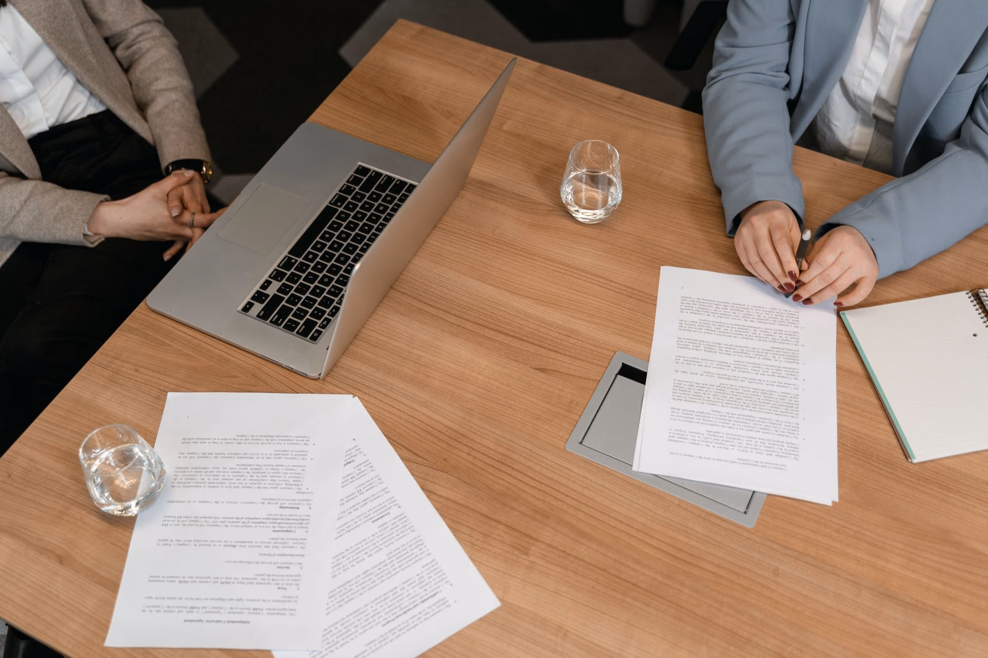 Two people sitting at a table with documents, a laptop, glasses of water, and a notebook.
