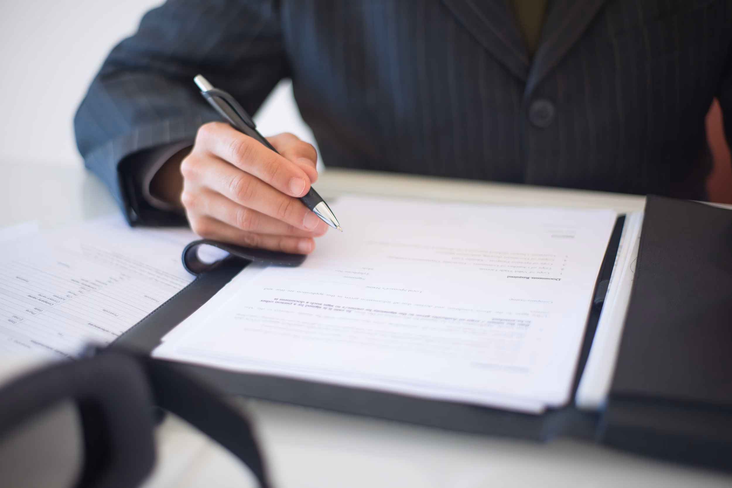 Person in business suit holding pen over documents on a clipboard, preparing to sign.