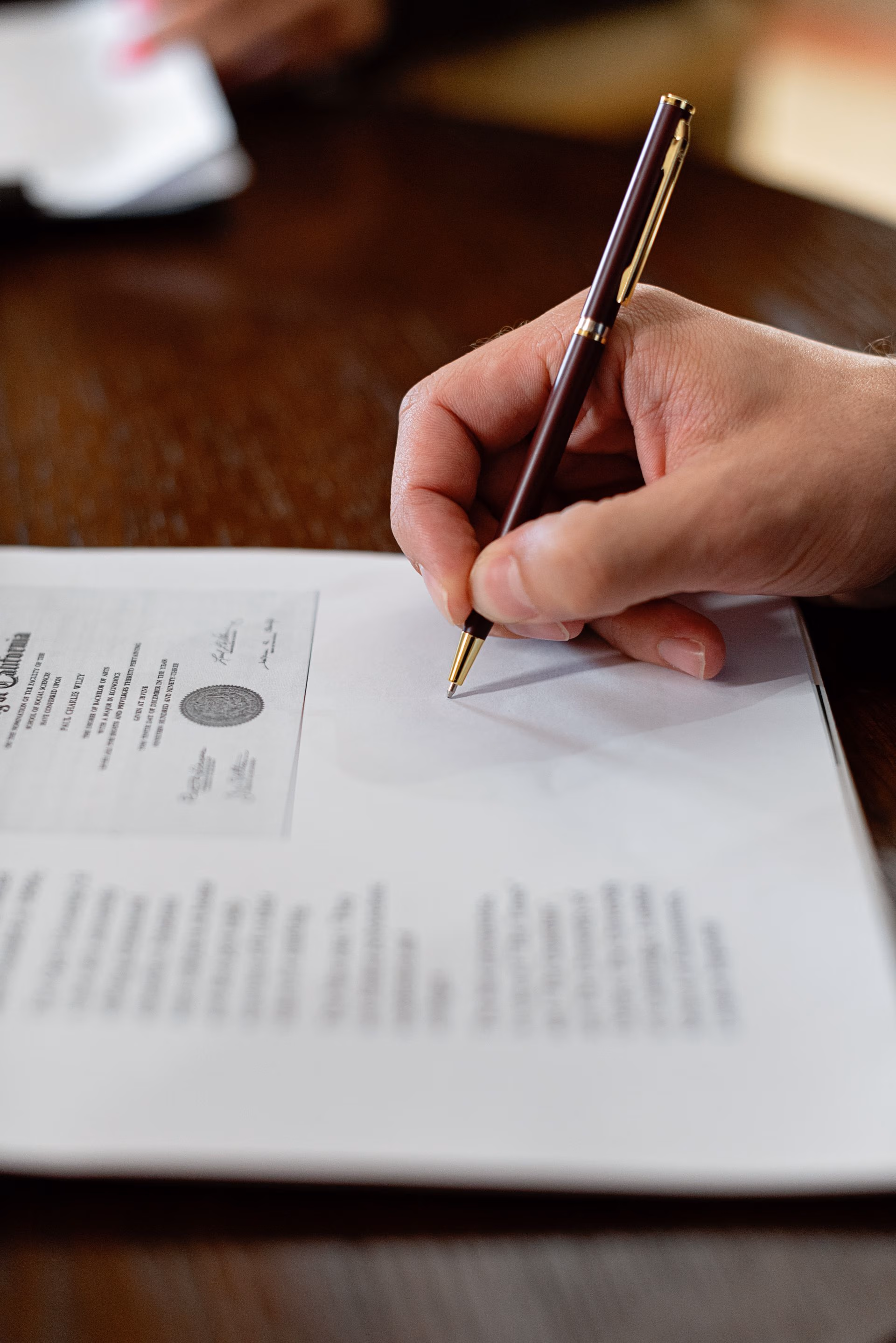 Close-up of a hand holding a pen poised to write on a paper document on a wooden table.