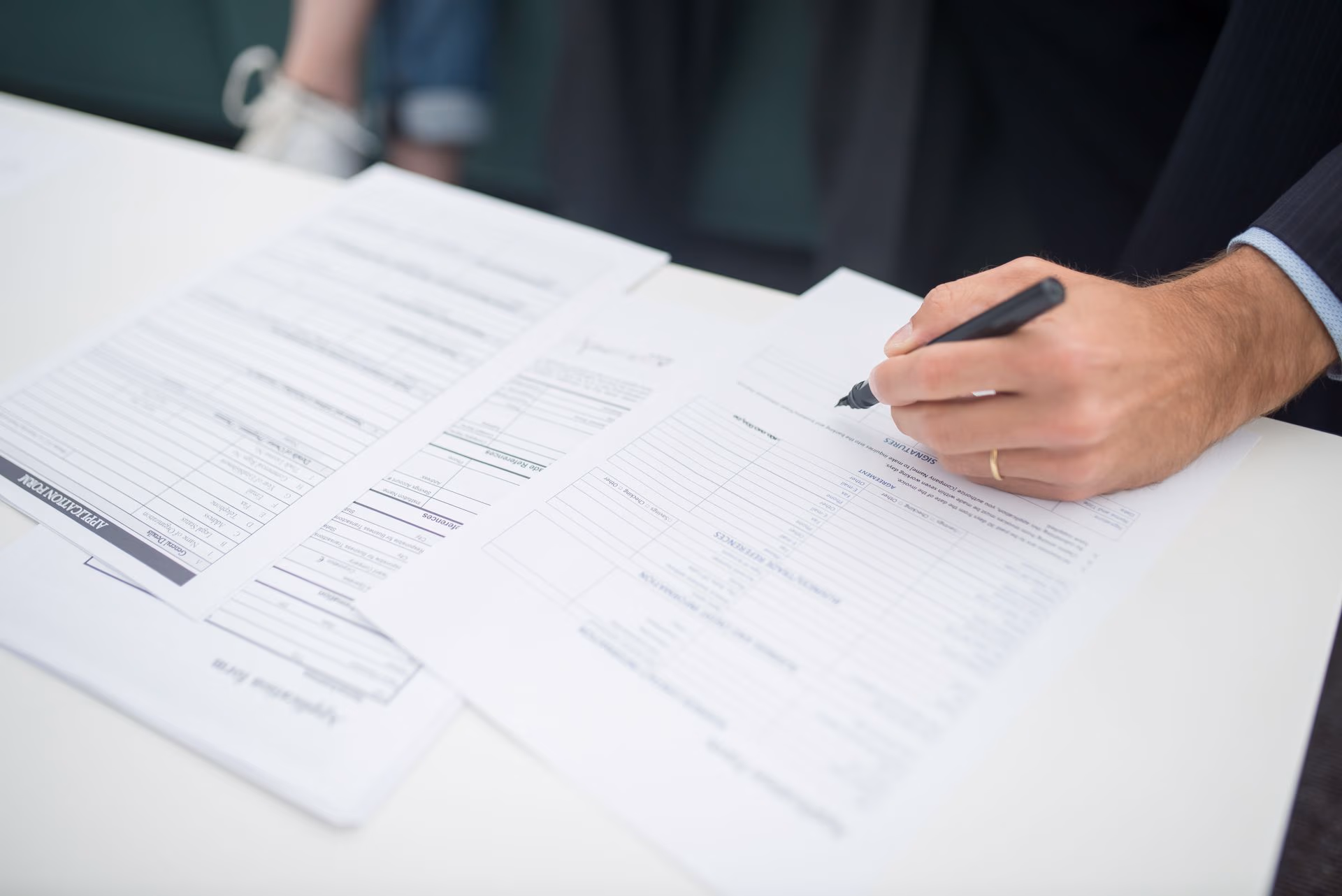 Person wearing a suit fills out a form with a pen on a white desk.