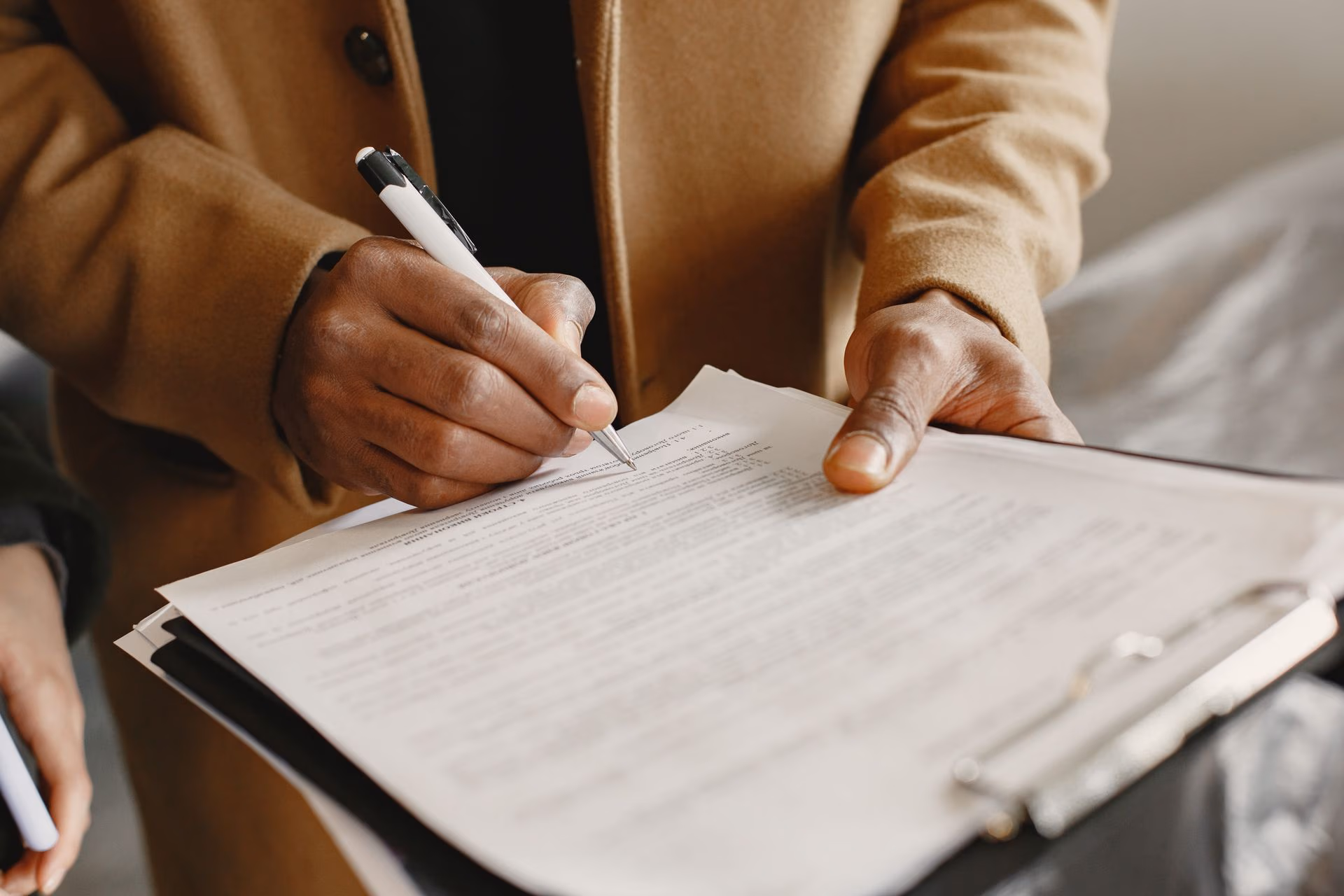 Person in brown coat signing a document on a clipboard with a pen.