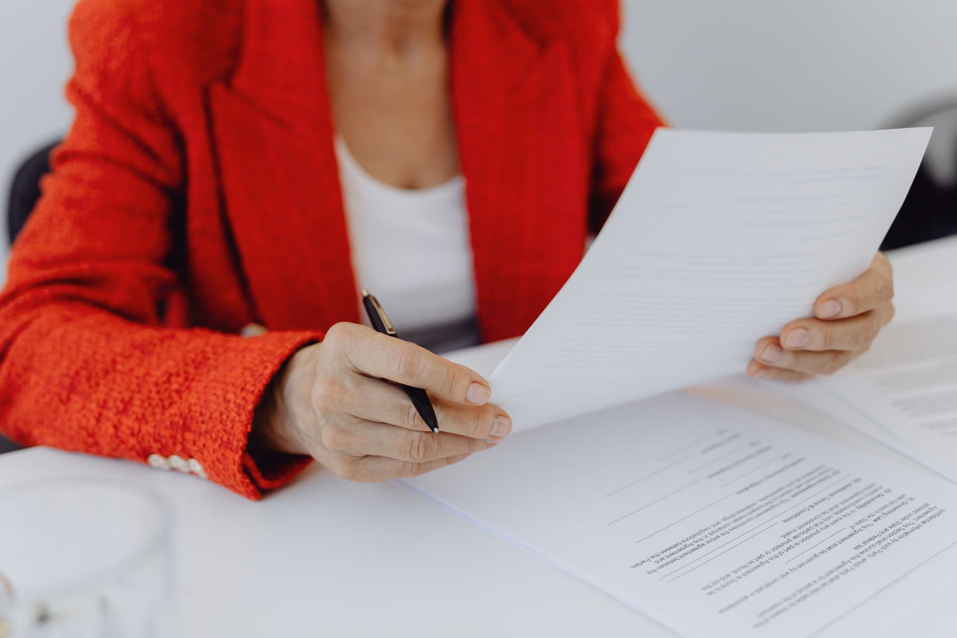 Person in red jacket holding and reviewing papers at a white desk with a pen.