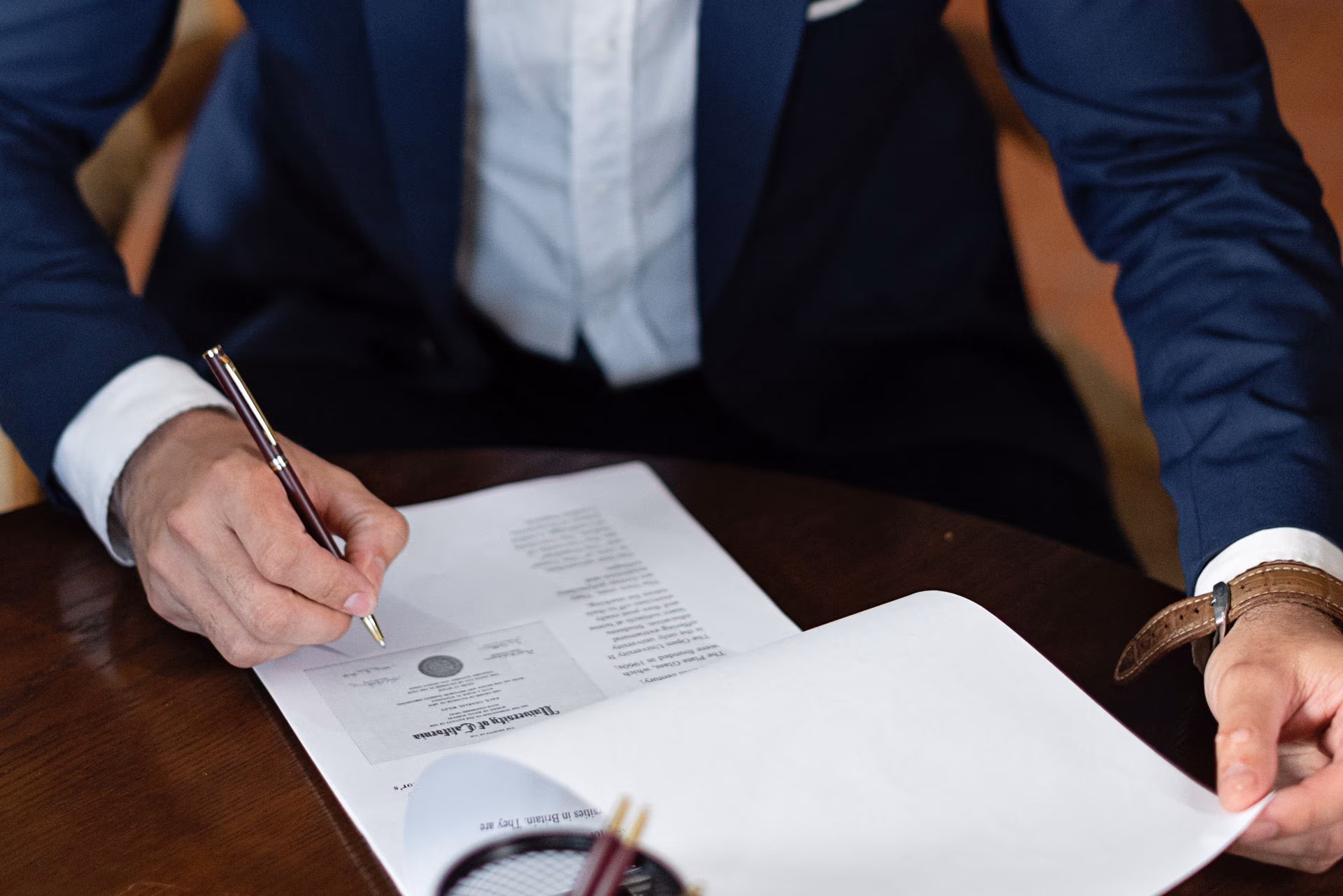 Person in suit signing a document with a pen at a wooden desk.