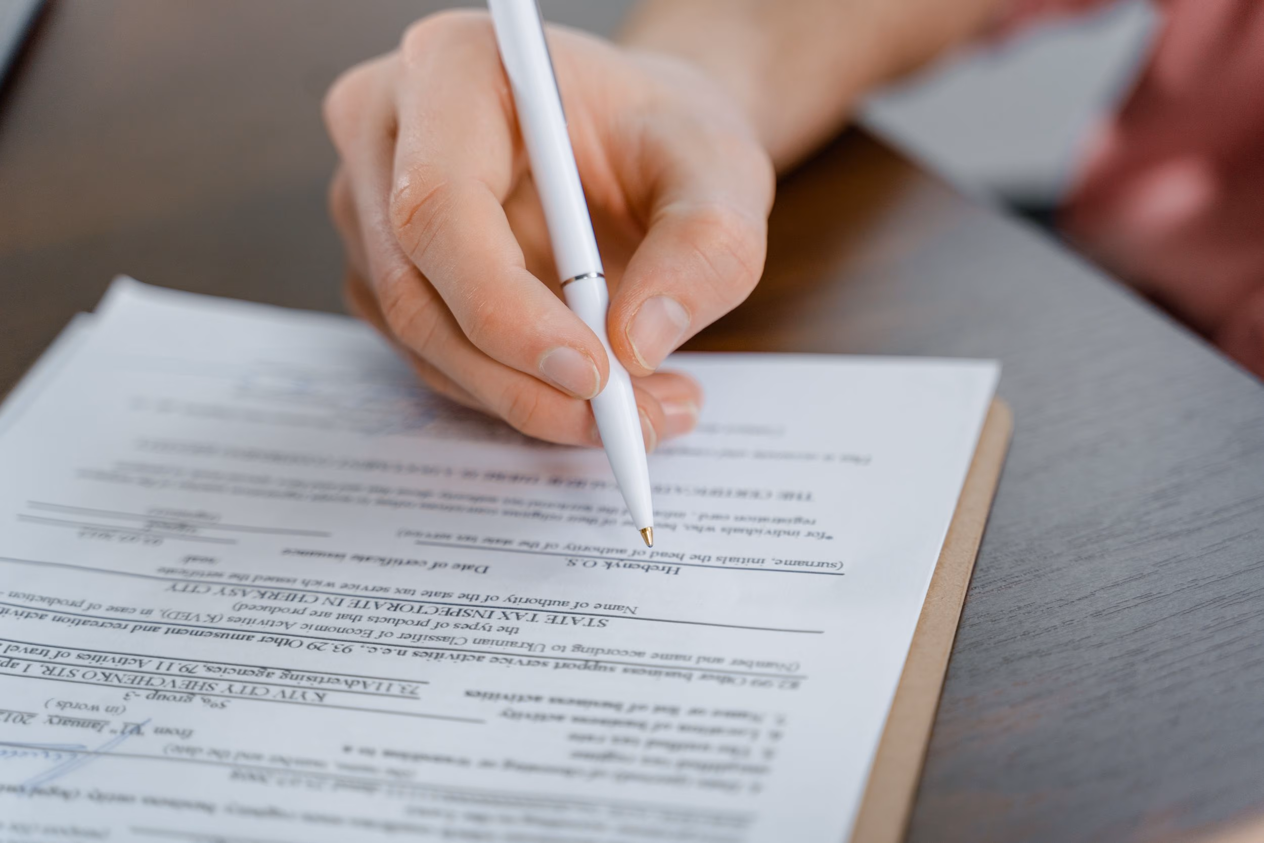 Hand holding a white pen poised above a document on a clipboard on a gray wooden table.