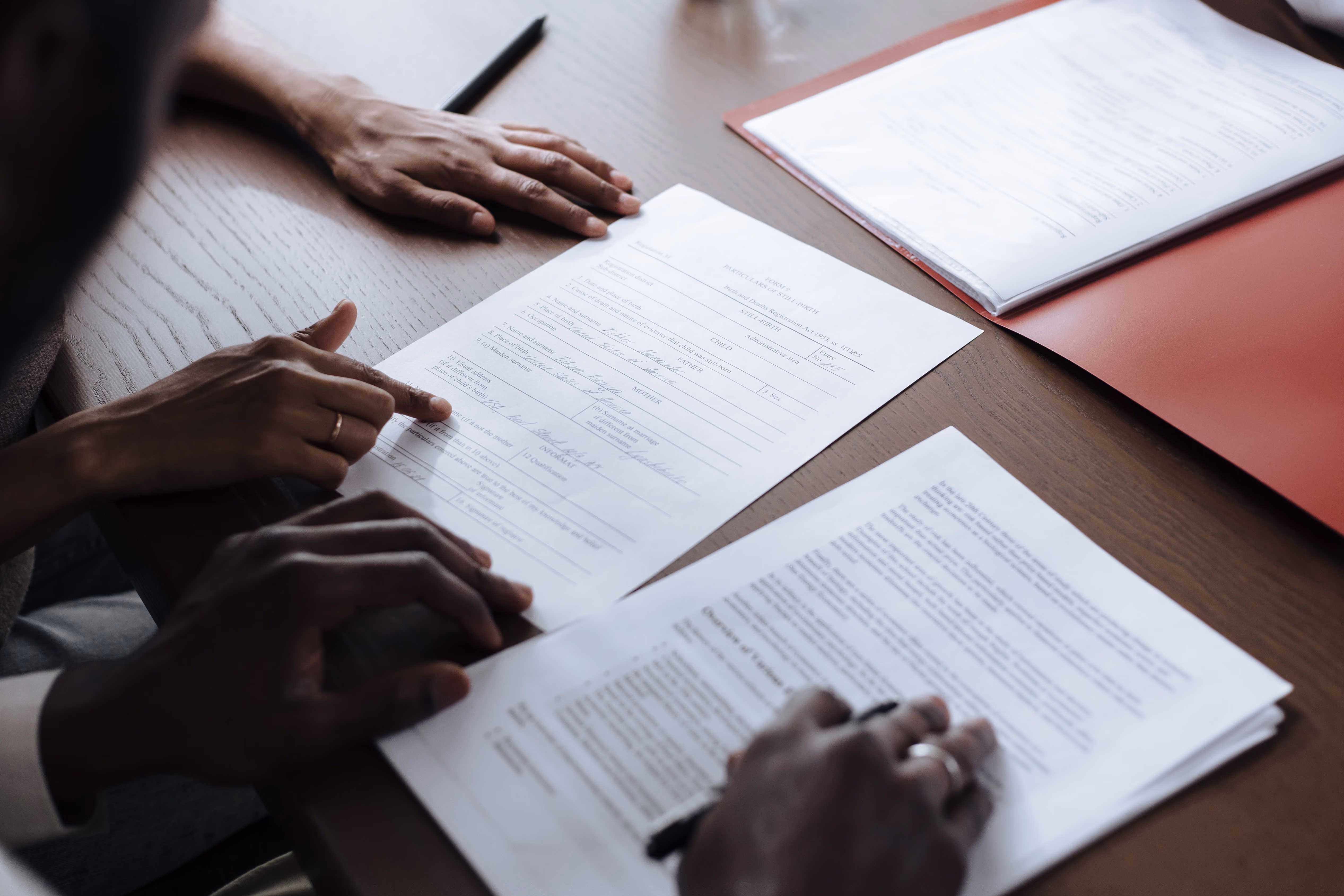 Two people reviewing and pointing at official documents on a wooden table, one holding a pen.