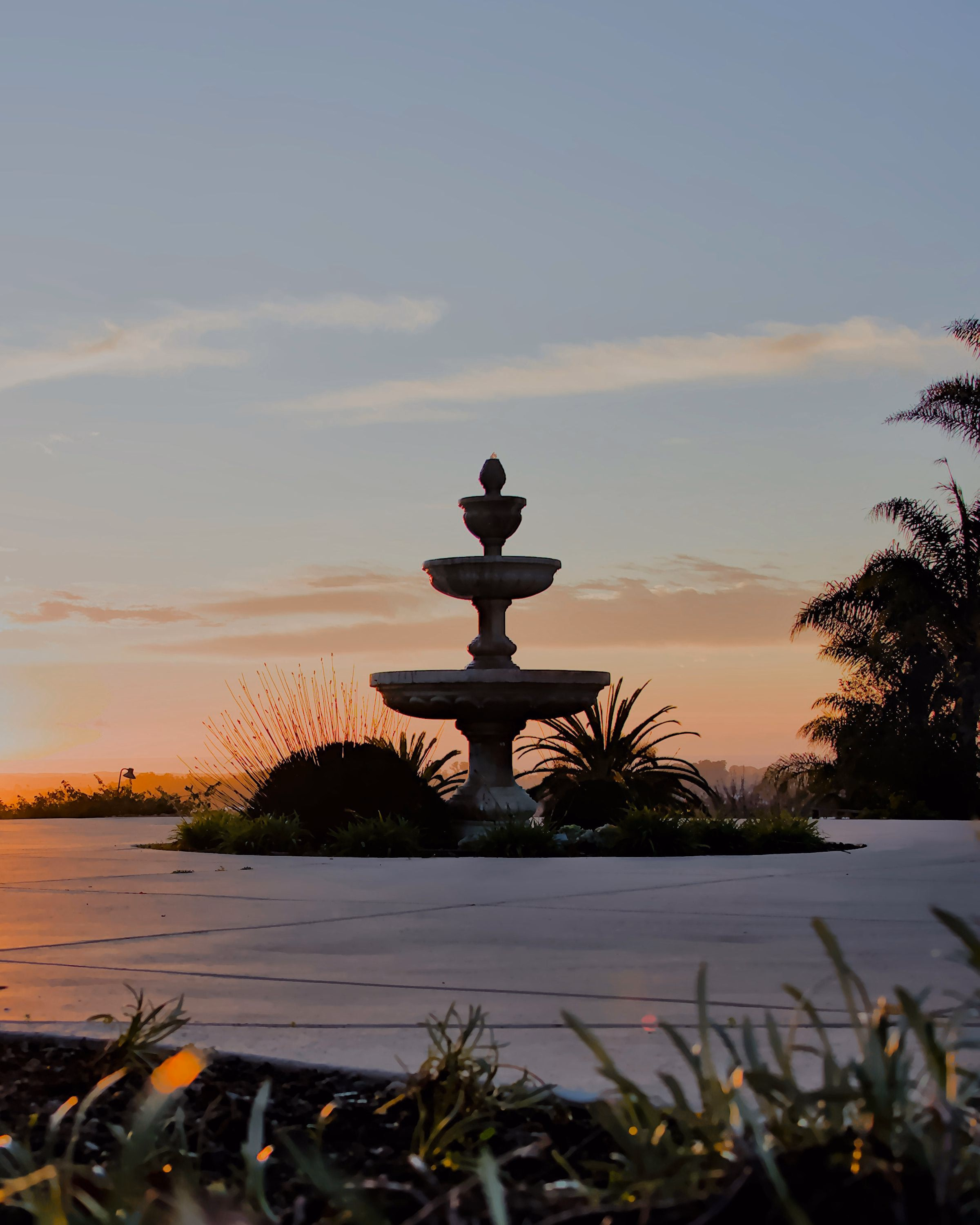 A tiered stone fountain on an open plaza at sunset, with palms and low hills on the horizon.