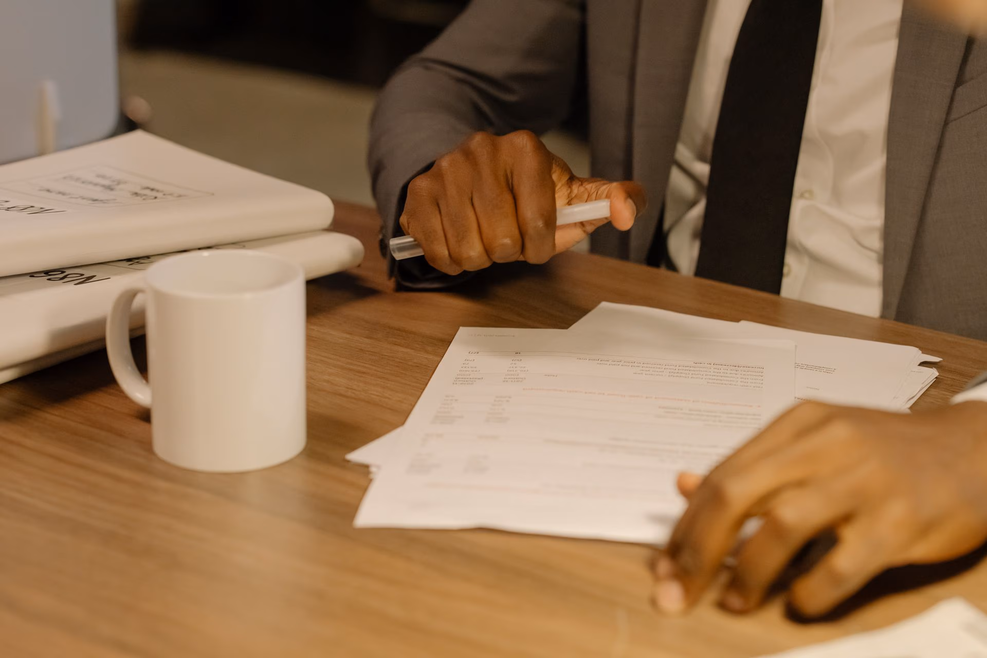 A person in suit holding a pen reviewing documents on a wooden table beside a white coffee mug.