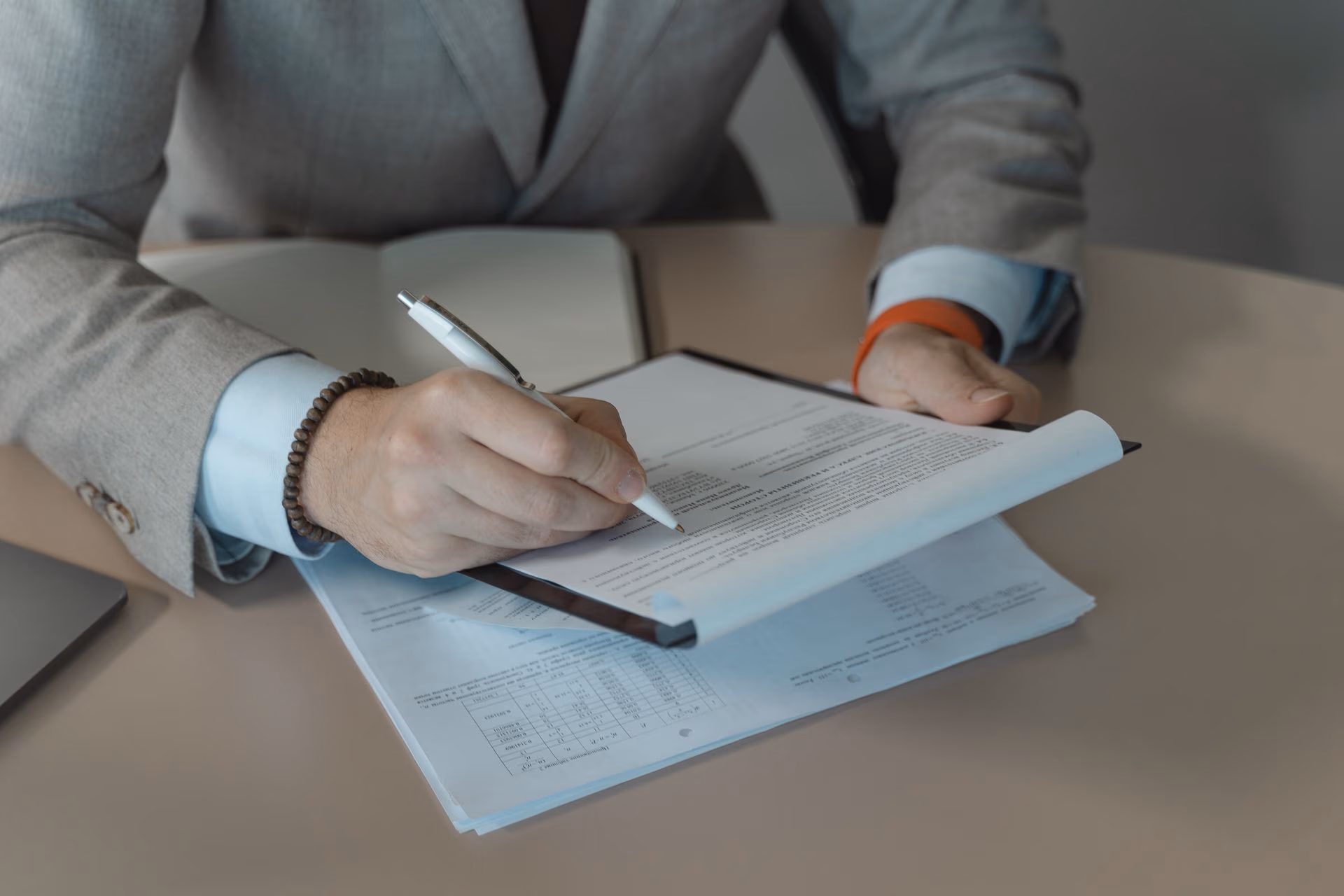Person in a gray suit holding a pen and reviewing documents on a clipboard at a table.