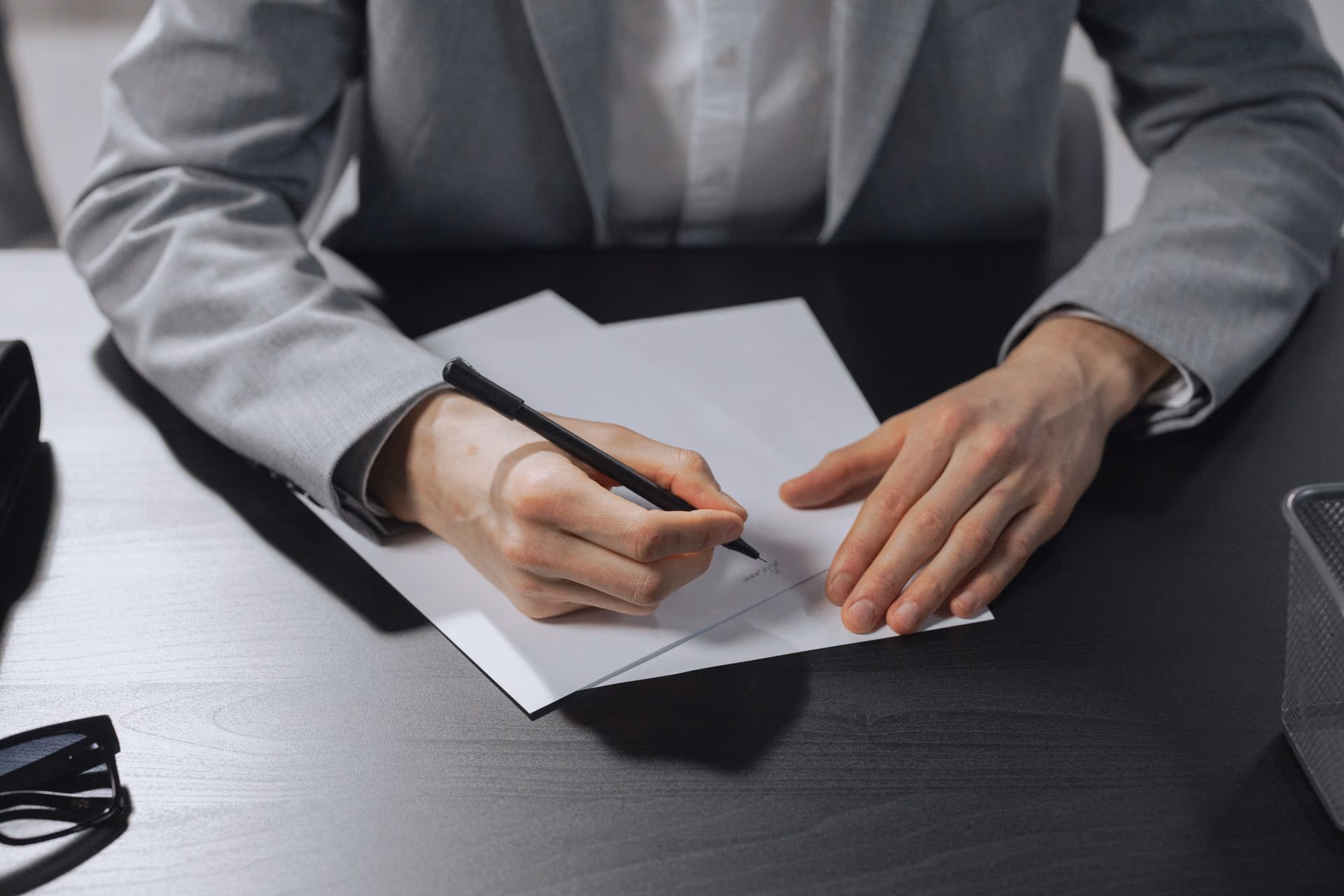 Person in gray suit writing on white papers with a black pen on a dark desk.